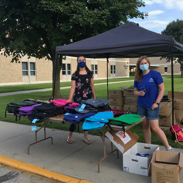 Two female school drive volunteers standing outside behind a table filled with backpacks